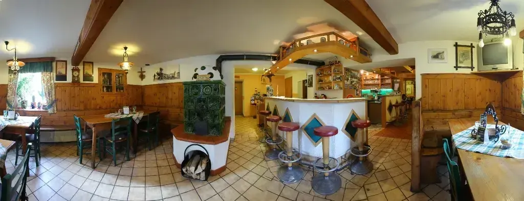 Ein gemütliches Restaurant mit Holzwänden, gefliestem Boden und einer Theke mit Hockern. Ein Holztisch mit Stühlen ist aufgestellt, und ein Holzschrank ist links platziert. Ein gerahmtes Bild ist an der Wand montiert. Ein grüner Ofen steht links, und eine Lampe ist an der Decke montiert.