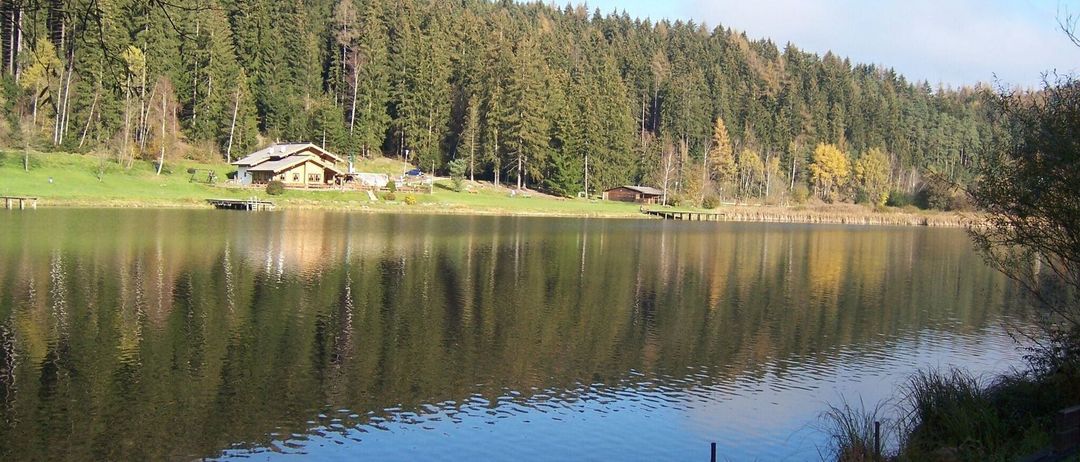 A serene lake surrounded by a lush green hillside with a house on the shore and a wooden dock extending into the water.
