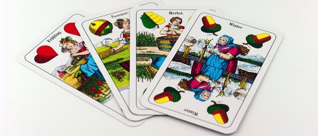 Vier Spielkarten liegen auf einer weißen Oberfläche. Jede Karte hat ein einzigartiges Design und Text. Die erste Karte zeigt eine Frau mit Hut und Korb. Die zweite Karte hat ein gelbes Blatt und eine Frau mit Korb. Die dritte Karte zeigt eine Frau mit Besen und Korb. Die vierte Karte zeigt eine Frau mit Hut und Korb.