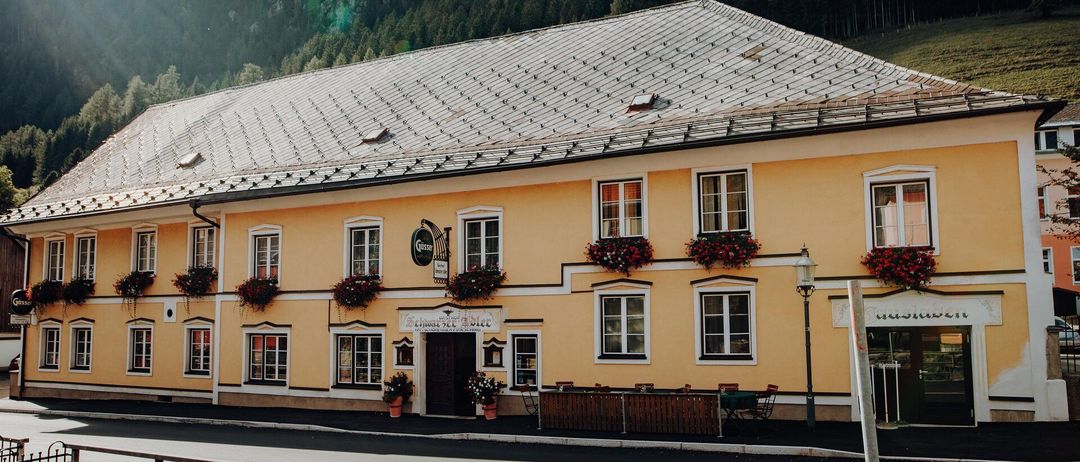 Ein gelbes Gebäude mit einem grauen Dach liegt am Fuße eines Berges, geschmückt mit roten Blumen an den Fenstern. Es hat ein Schild, das auf ein Restaurant hinweist, und eine Holzumzäunung mit Stühlen und Tischen draußen.