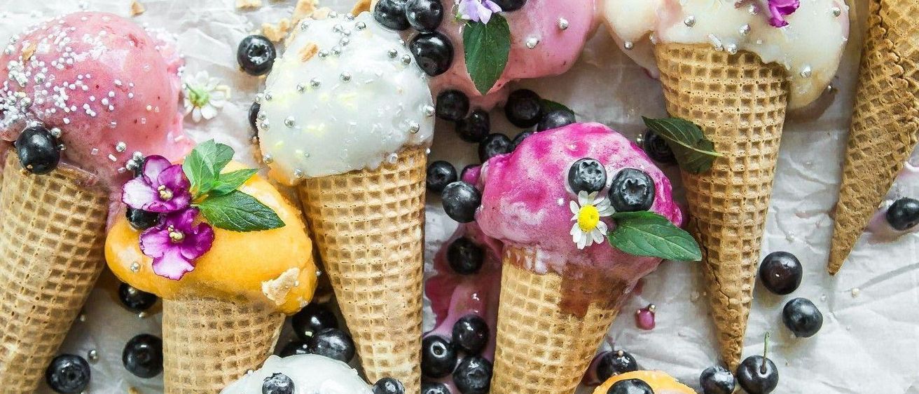 Eine Vielzahl von Eiswaffeln ist auf einem weißen Hintergrund angeordnet, jede mit verschiedenen Früchten und Blumen verziert. Die Kegel umfassen rosa, gelbe und weiße Eiscreme, wobei Beeren und Minzblätter zur Dekoration beitragen.