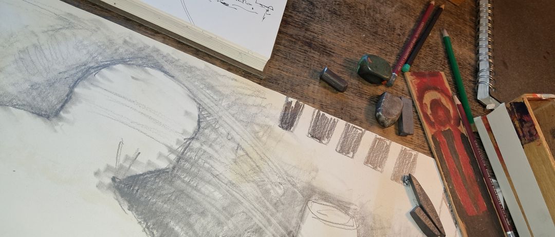 Ein Kunstarbeitsplatz mit verschiedenen Werkzeugen. Eine Skizze einer Brücke auf einem Papier. Bleistifte, ein Malkasten und ein Marker sind auf dem Schreibtisch.