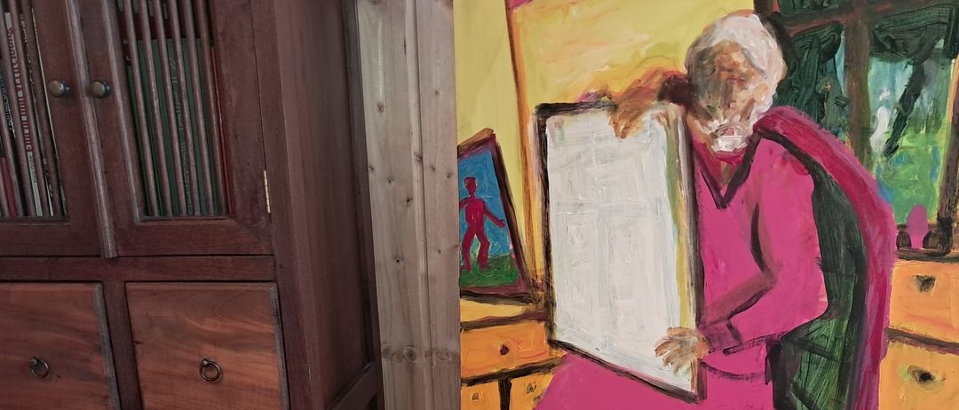 Eine ältere Person sitzt auf einem Stuhl in einem Raum mit einem Holzschrank und einem Gemälde an der Wand. Das Gemälde zeigt eine Person, die ein weißes Brett hält. Der Boden ist aus Holz.