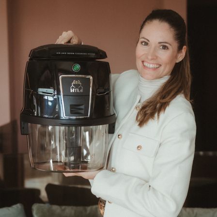 Eine lächelnde Frau in einem weißen Pullover und einer braunen Hose hält ein schwarzes Hylaa Luft- und Raumreinigungssystem in einem Raum mit einer rosa Wand und einer grünen Couch.