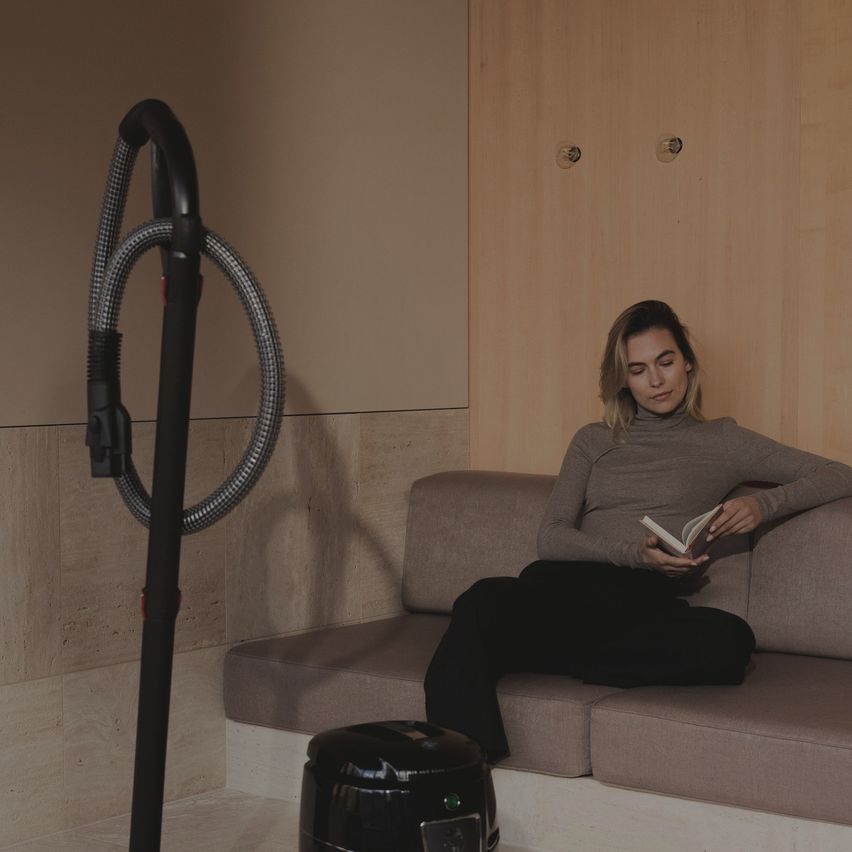 Eine Frau in einem grauen Rollkragenpullover sitzt auf einer Couch und liest ein Buch. Ein Staubsauger steht neben ihr.