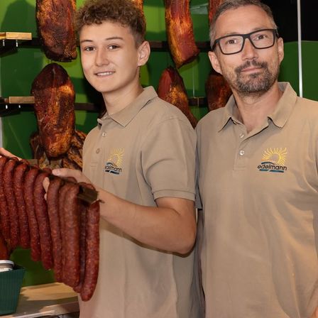 Zwei Männer in passenden Hemden stehen neben einem Regal mit gepökeltem Fleisch. Der Mann auf der linken Seite hält eine Handvoll Würste und lächelt. Der Mann auf der rechten Seite trägt eine Brille.