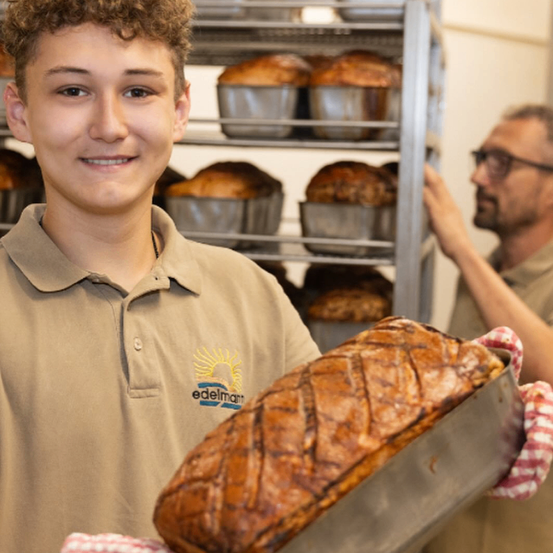 Ein junger Mann in einem Poloshirt hält einen Braten mit knuspriger brauner Außenseite. Er hat ein Lächeln im Gesicht und steht vor einem Regal mit mehreren Broten in Silberschalen.