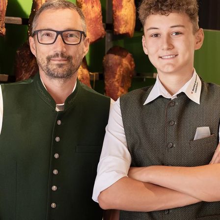 Zwei Männer stehen nebeneinander vor einer grünen Wand. Der Mann auf der linken Seite trägt eine Brille und eine grüne Weste, während der Mann auf der rechten Seite ein weißes Hemd und eine grüne Weste trägt.