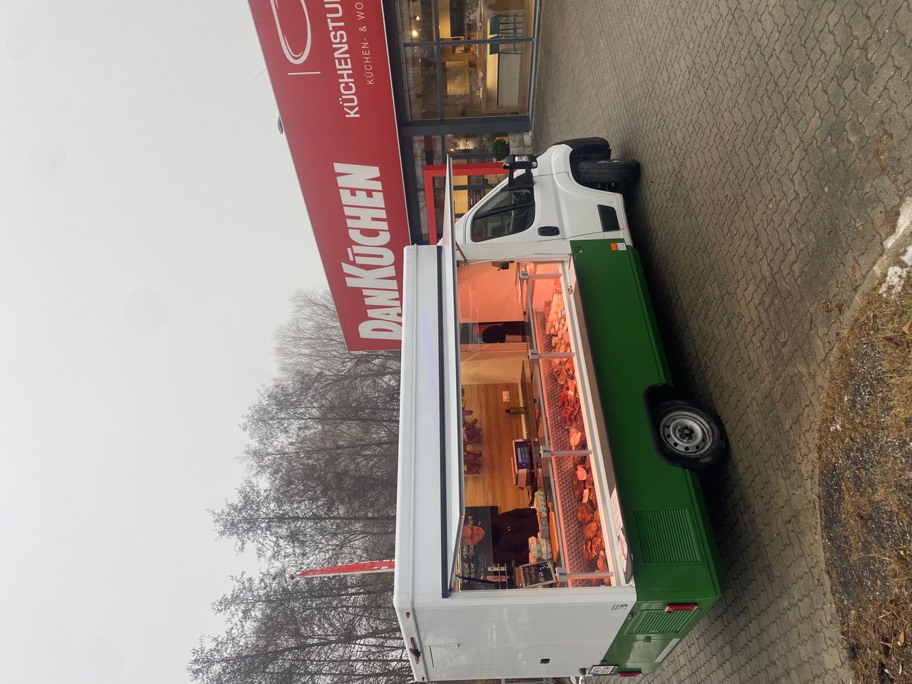 Ein grüner und weißer Metzgerwagen vor einem Geschäft namens Dankechen. Der Himmel ist bedeckt.