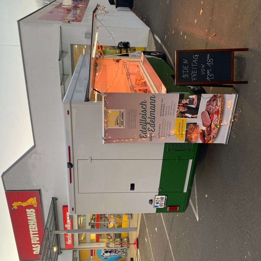 Ein Imbisswagen ist vor einem Geschäft geparkt und zeigt ein Banner mit einem Fleischteller und einem Preisschild. Das Geschäft hat ein rotes Schild mit einem Hund und dem Wort "Futterhaus".
