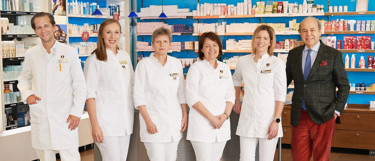 Eine Gruppe von Apothekern in weißen Uniformen steht hinter einer Apothekentheke, lächelt und posiert für ein Foto mit einem Mann im Anzug und Krawatte.