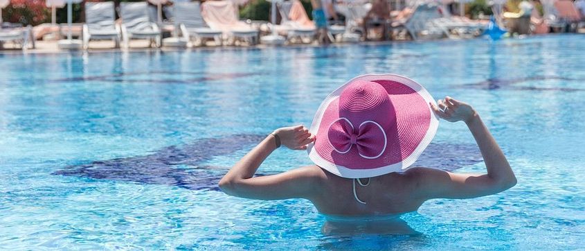Eine Frau in einem rosa Hut schwimmt in einem Pool in einem Resort mit mehreren Sonnenschirmen und Liegestühlen im Hintergrund.