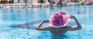 Eine Frau in einem rosa Hut schwimmt in einem Pool in einem Resort mit mehreren Sonnenschirmen und Liegestühlen im Hintergrund.