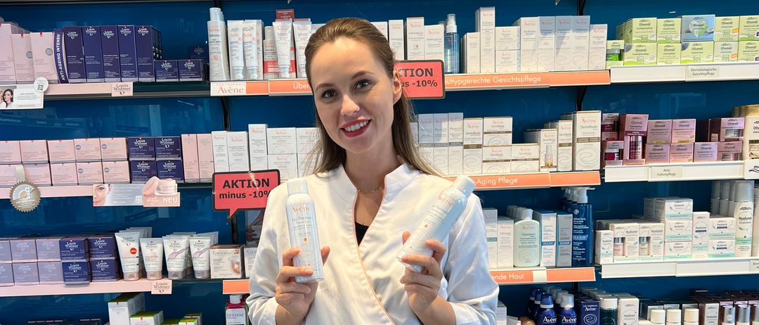 Eine Frau in einem weißen Gewand lächelt und posiert mit zwei Flaschen in einer Apotheke, umgeben von Regalen mit verschiedenen Hautpflegeprodukten.