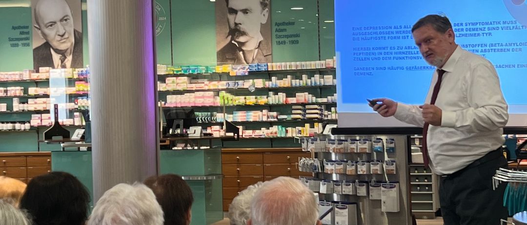 Ein Mann in weißem Hemd und Krawatte hält eine Präsentation in einem Raum mit einer Projektionsleinwand. Mehrere Menschen sitzen vor ihm in Stühlen.