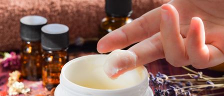 Eine Hand taucht den Finger in eine Creme-Schale, mit Lavendel und ätherischen Ölen im Hintergrund.