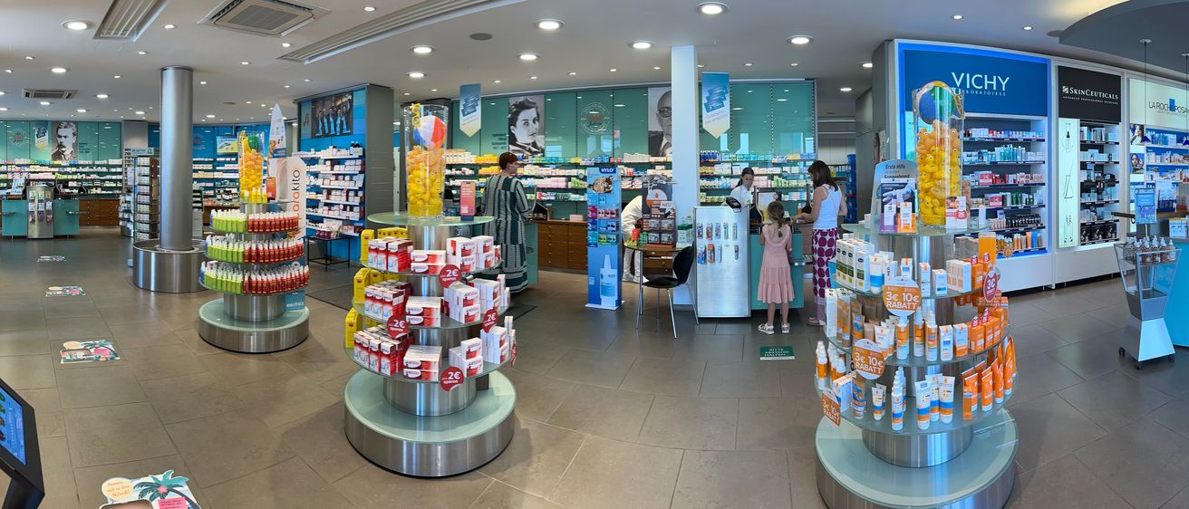 Im Inneren einer Apotheke interagieren Kunden und Mitarbeiter. Eine Frau am Tresen hilft einem Mädchen, während eine andere Frau in der Nähe steht. Regale mit Produkten und eine Display mit Süßigkeiten verleihen dem Geschäft Atmosphäre.