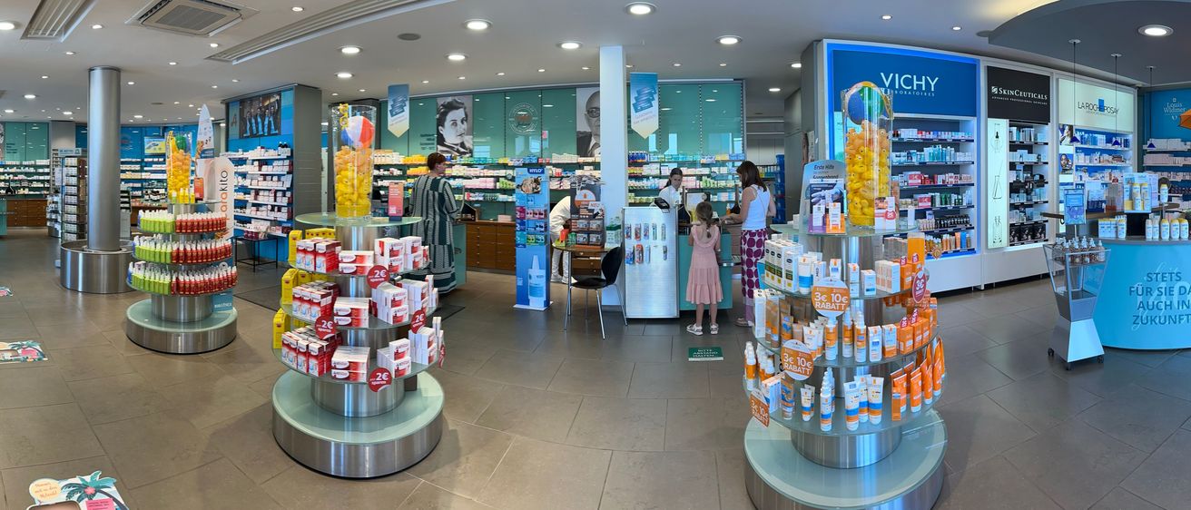 Im Inneren einer Apotheke interagieren Kunden und Mitarbeiter. Eine Frau am Tresen hilft einem Mädchen, während eine andere Frau in der Nähe steht. Regale mit Produkten und eine Display mit Süßigkeiten verleihen dem Geschäft Atmosphäre.