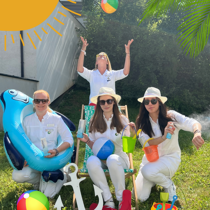 Drei Frauen in weißen Kleidern posieren auf einem Rasen. Eine Frau kniet und hält einen Eimer, eine andere sitzt in einem Stuhl und hält einen Ball, und die dritte steht hinter ihnen und schaut auf einen Ball in der Luft.