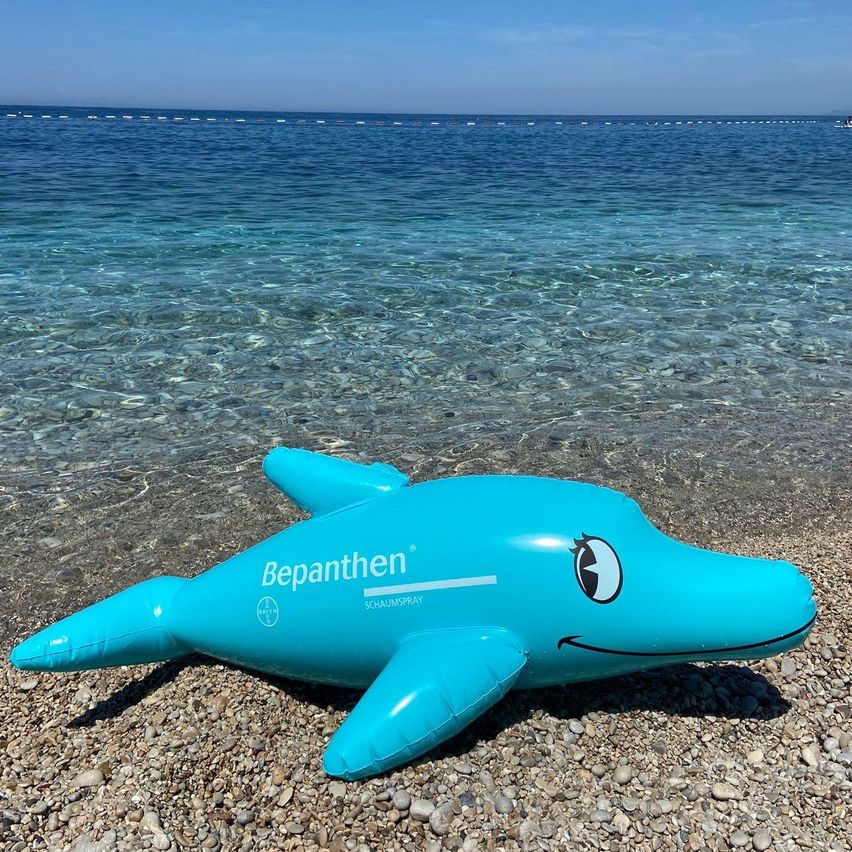 Ein großer blauer aufblasbarer Delfin, mit Bepanthen-Marke, liegt am Felsstrand am Meer. Das Wasser ist ruhig mit leichten Wellen.