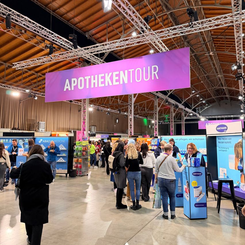 Eine belebte Indoor-Veranstaltung mit Menschen, die sich um Stände versammeln. Ein großes Schild mit der Aufschrift 'APOTHEKENTOUR' hängt von der Decke. Besucher interagieren mit den Ständen, einige stehen und andere gehen herum. Es gibt mehrere Tische mit ausgestellten Produkten.