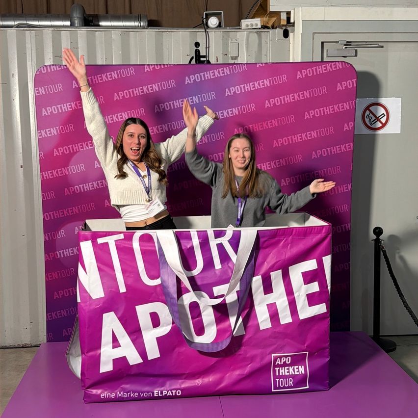 Zwei Frauen sind in einer großen lila Tasche, auf der 'Apotheken Tour' steht. Sie lächeln und heben ihre Hände. Sie tragen Ausweise um den Hals. An der Wand befindet sich ein Schild mit der Aufschrift 'Rauchen verboten'.