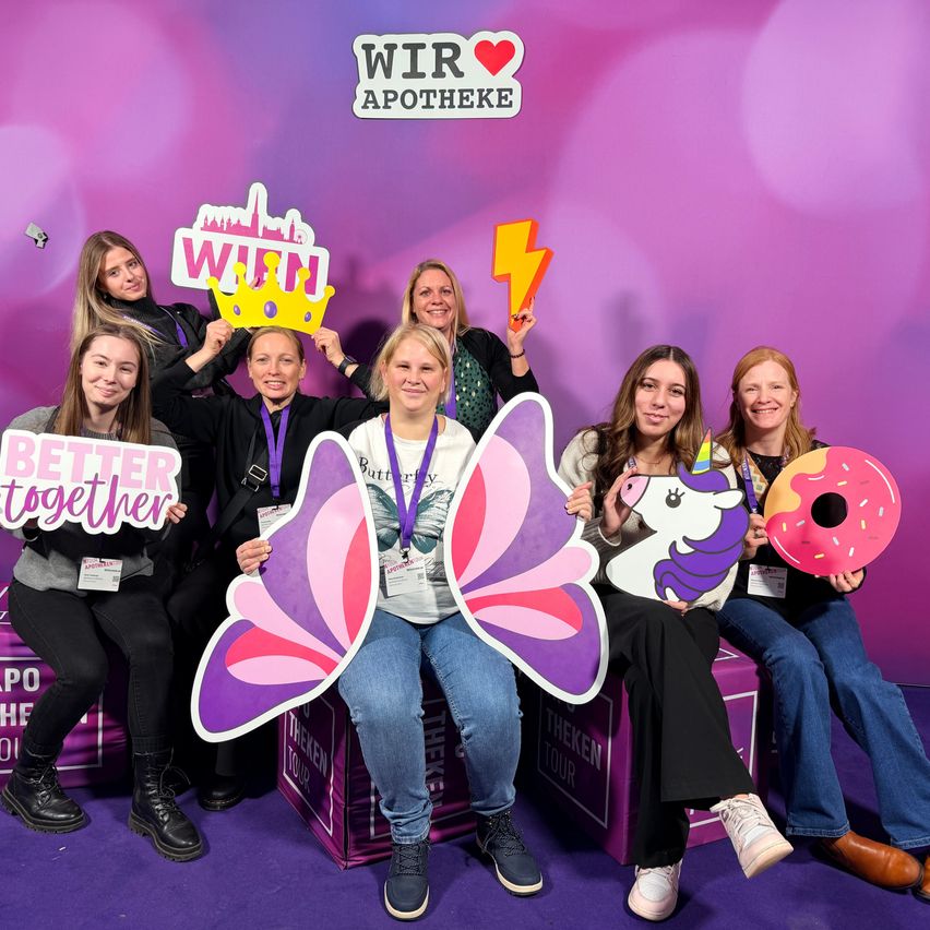 Eine Gruppe von Frauen in lila und schwarz posiert für ein Foto mit Requisiten wie einer Krone, Schildern und einem Einhorn-Ausschnitt vor einem lila Hintergrund mit dem Wort Apotheke darauf.