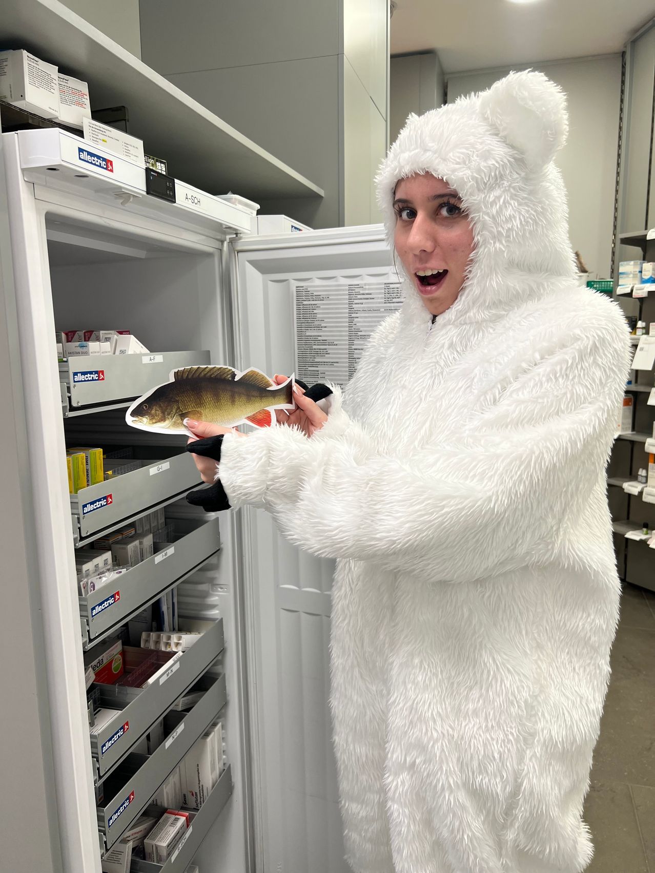 Eine Person in einem weißen Bärenkostüm hält eine Fischausschnittfigur in einem Kühlschrank mit beschrifteten Regalen.