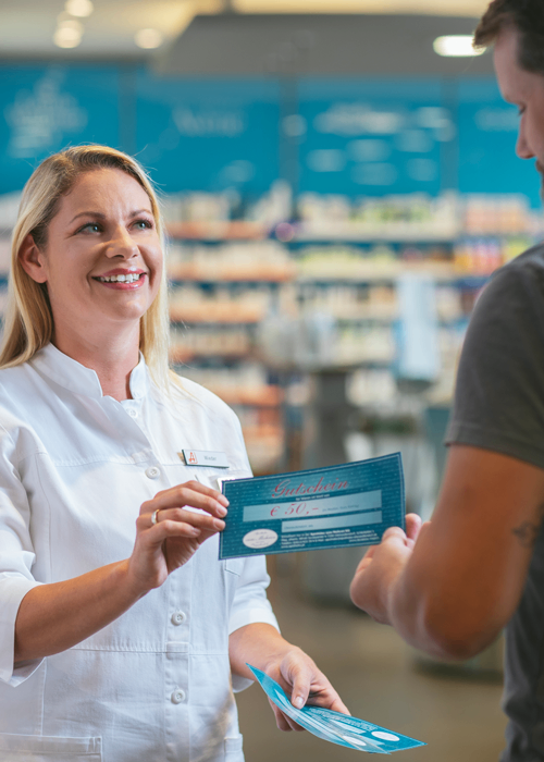 Eine lächelnde Frau in einer weißen Uniform hält einen Gutschein und ein blaues Stück Papier in der Hand, steht in einer Apotheke und spricht mit einem Mann, der ein graues T-Shirt trägt.