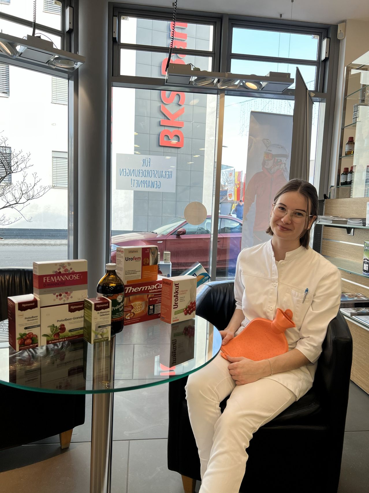 Eine Frau in einem weißen Laborkittel sitzt lächelnd in einem Stuhl im Geschäft, mit einem Wärmflasche auf dem Schoß. Verschiedene Produkte stehen auf dem Tisch vor ihr. Das Fenster zeigt ein Gebäude und einen geparkten Wagen.