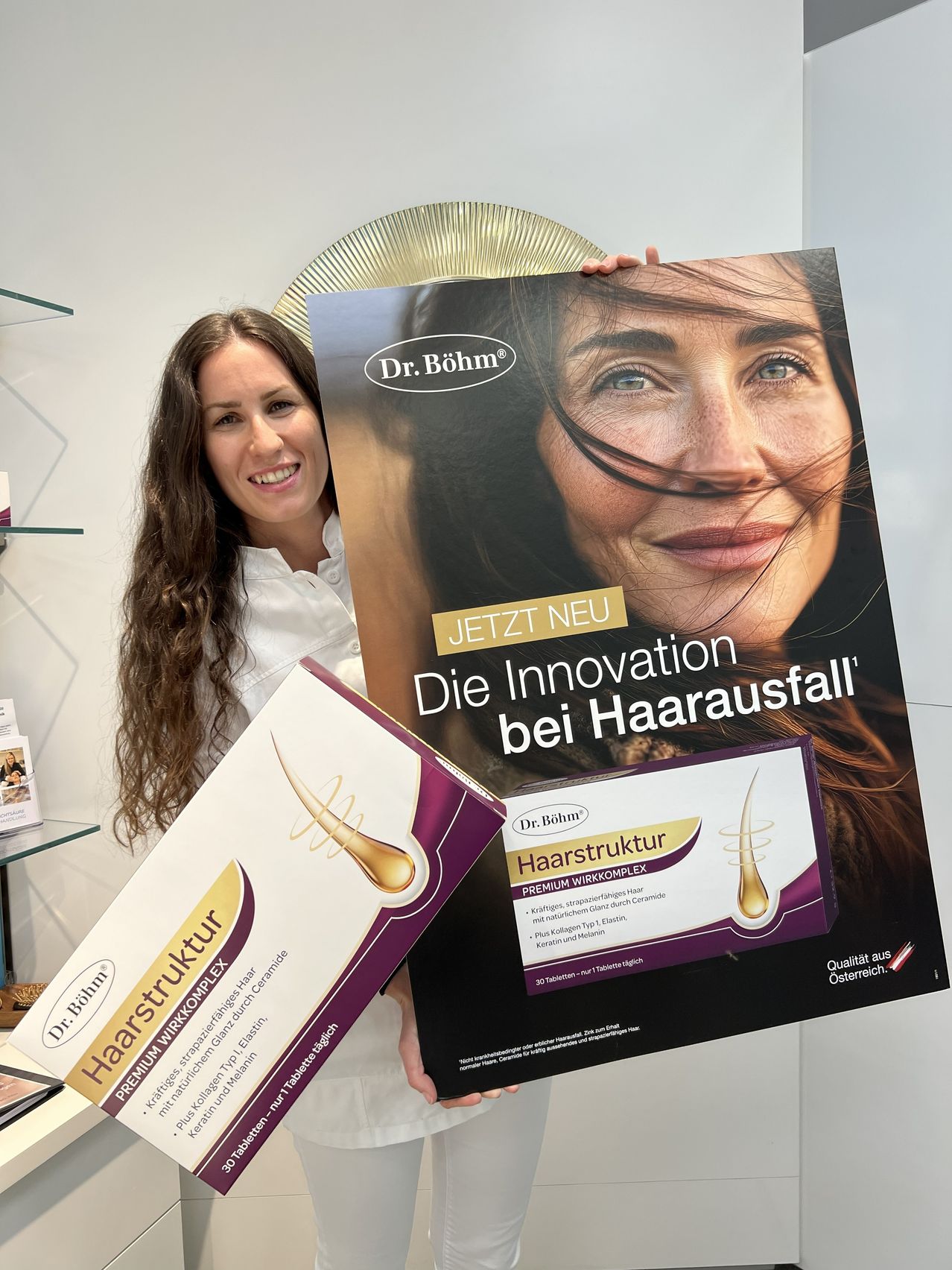 Eine Frau hält ein großes Plakat und eine Box mit Haarpflegeprodukten. Das Plakat besagt 'JETZT NEU Die Innovation bei Haarausfall'. Die Produktbox ist mit 'Haarstruktur' beschriftet.