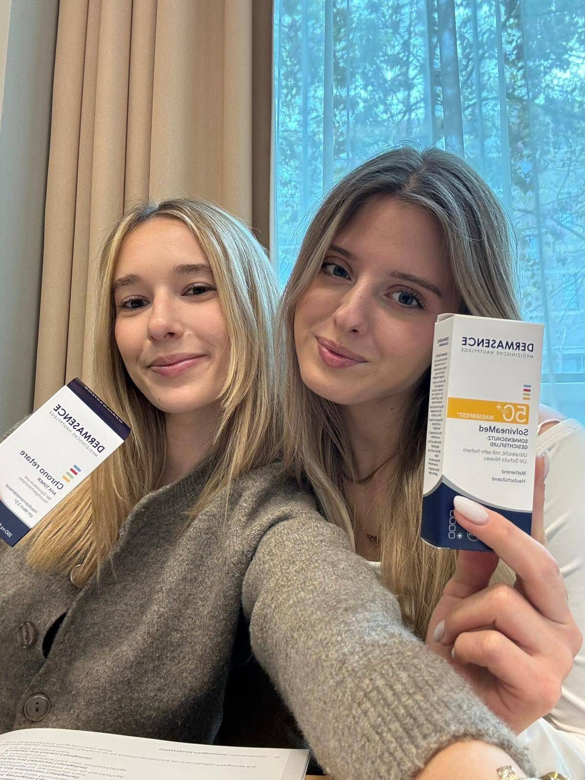 Zwei Frauen mit blonden Haaren lächeln und halten eine Box von Derma E. Sie haben blauen Nagellack.