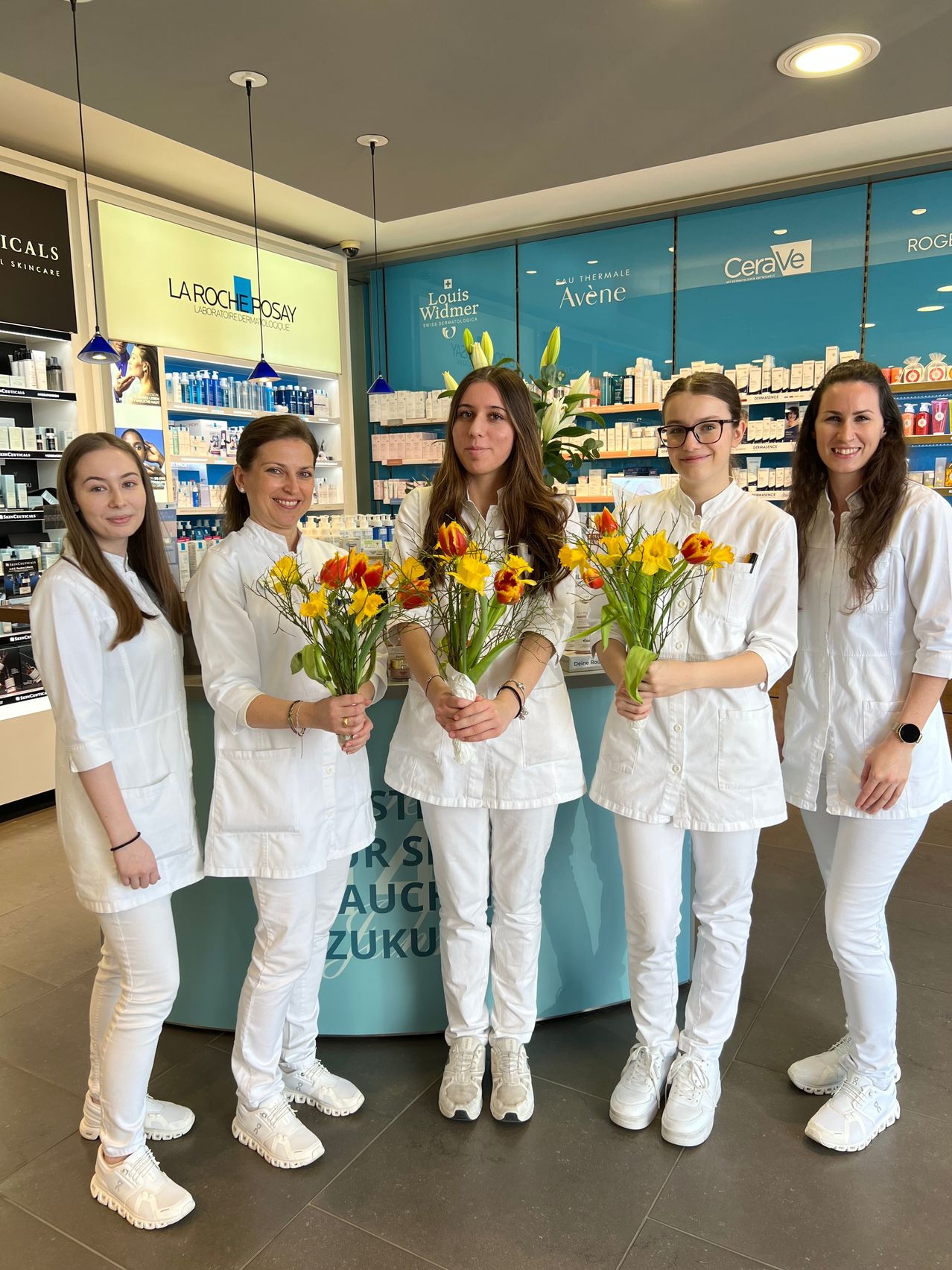 Five women in white lab coats stand in a beauty salon, smiling and holding bouquets of flowers. Behind them, a counter displays products. Advertisements for La Roche-Posay, CeraVe, and Roger & Gallet are visible.