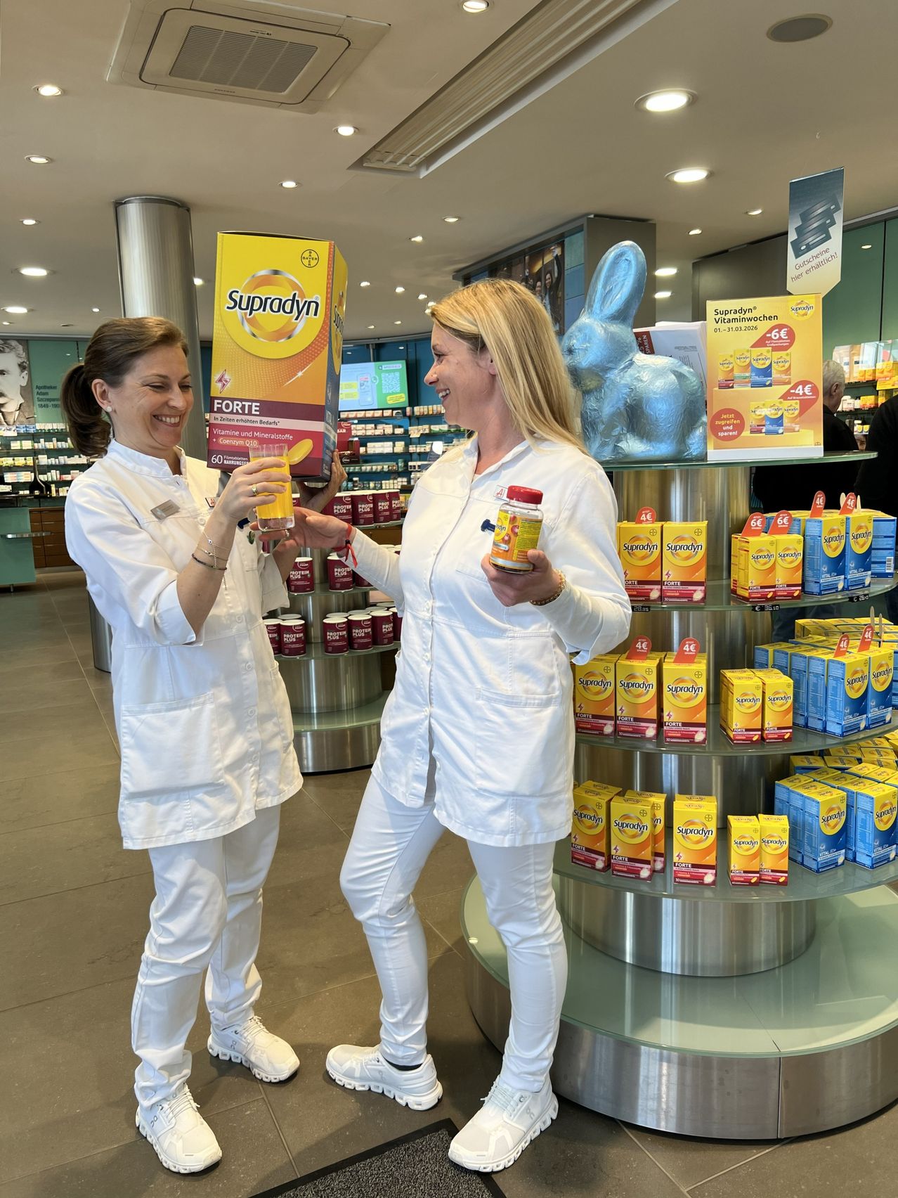 Zwei Frauen in weißen Uniformen befinden sich in einer Apotheke, lächeln und halten eine Flasche und ein Glas Saft. Dahinter steht eine Ausstellung von Supradyn-Produkten.