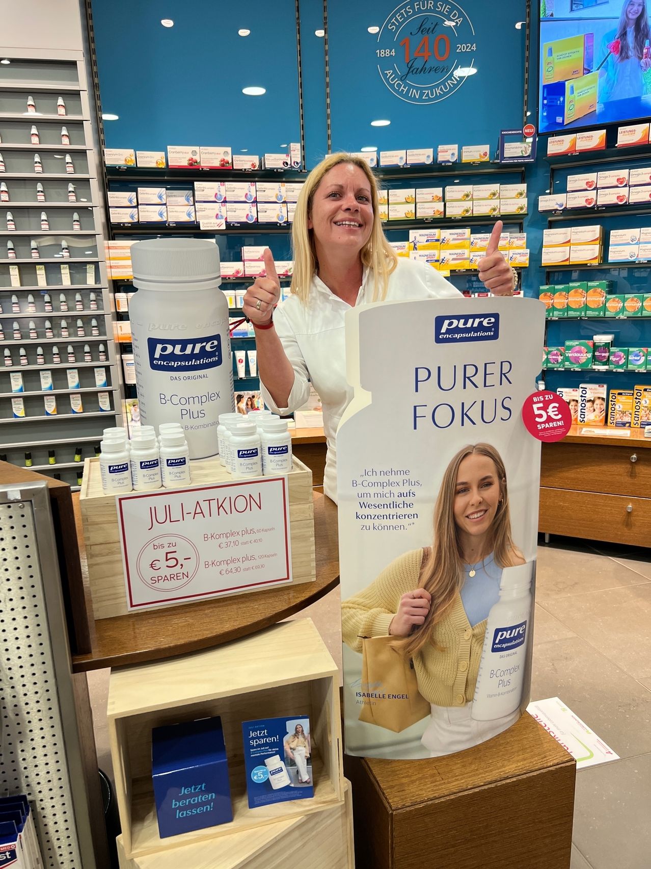 Eine Frau in einer Apotheke, die ein Schild hält und lächelt, mit einer Ausstellung von B-Complex Plus Nahrungsergänzungsmitteln hinter ihr.
