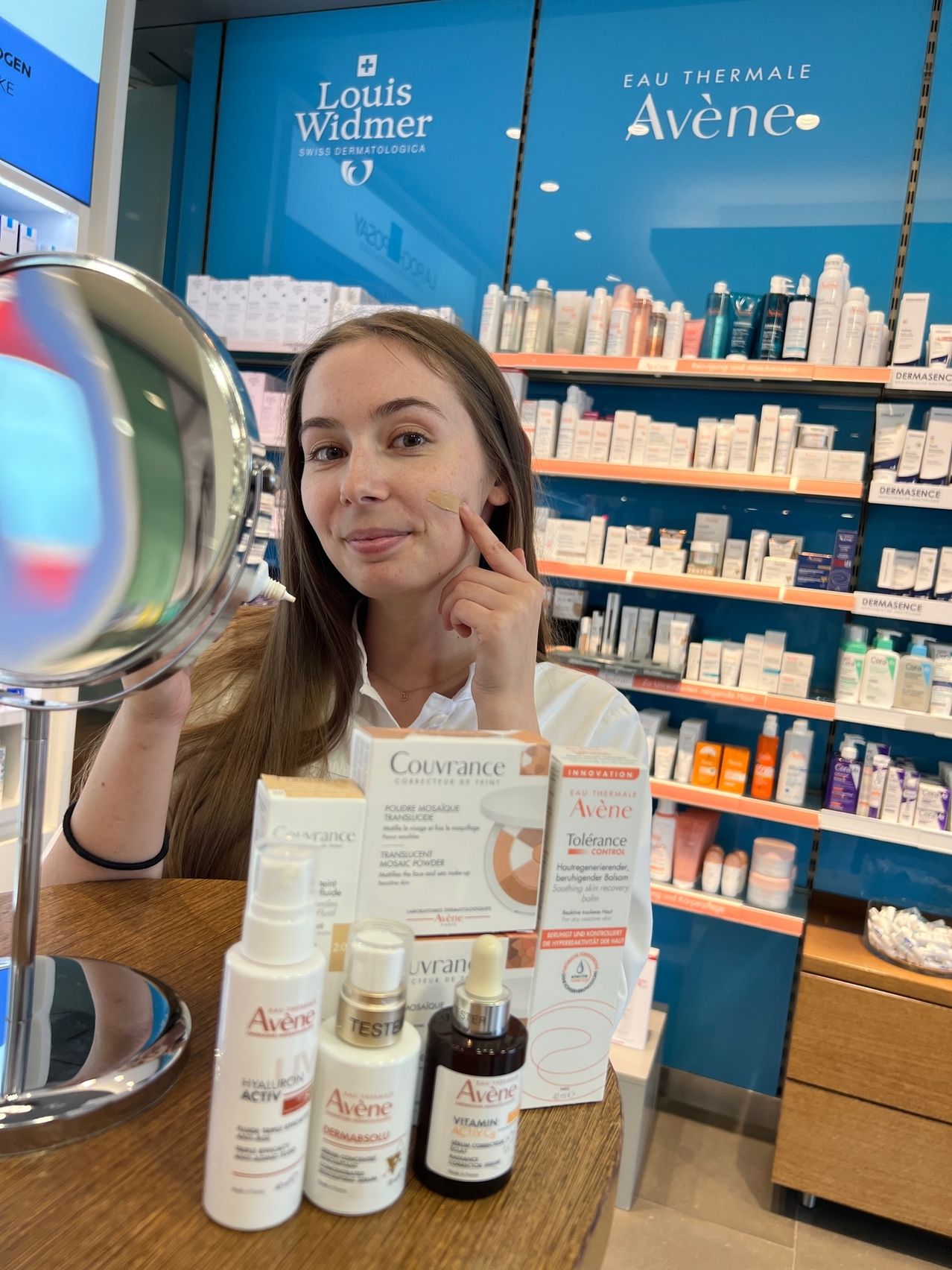 Eine Frau ist in einem Geschäft und trägt ein Kosmetikprodukt auf ihr Gesicht auf, während sie in einen Spiegel schaut. Verschiedene Hautpflegeprodukte stehen auf dem Tisch vor ihr.