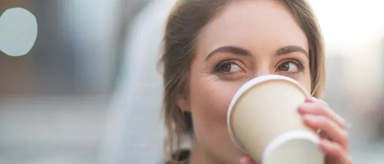 Eine Frau trinkt aus einem weißen Becher mit ernster Miene. Der Hintergrund ist unscharf.