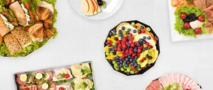 Eine Auswahl an Speisen, darunter Sandwiches, Obst und Fleisch, auf einer weißen Oberfläche. Teller und Schüsseln mit verschiedenen Speisen sind angeordnet.