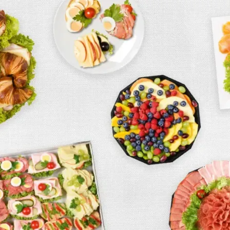 Eine Auswahl an Speisen auf Tellern, darunter Sandwiches, Obst und Fleisch, auf einer weißen Oberfläche angeordnet.