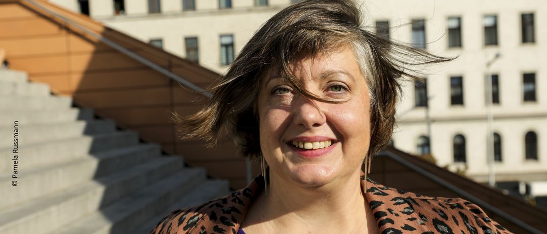 Eine Frau mit kurzen Haaren und einem Leopardenmuster-Jacke steht vor einem Gebäude mit vielen Fenstern und lächelt in die Kamera.
