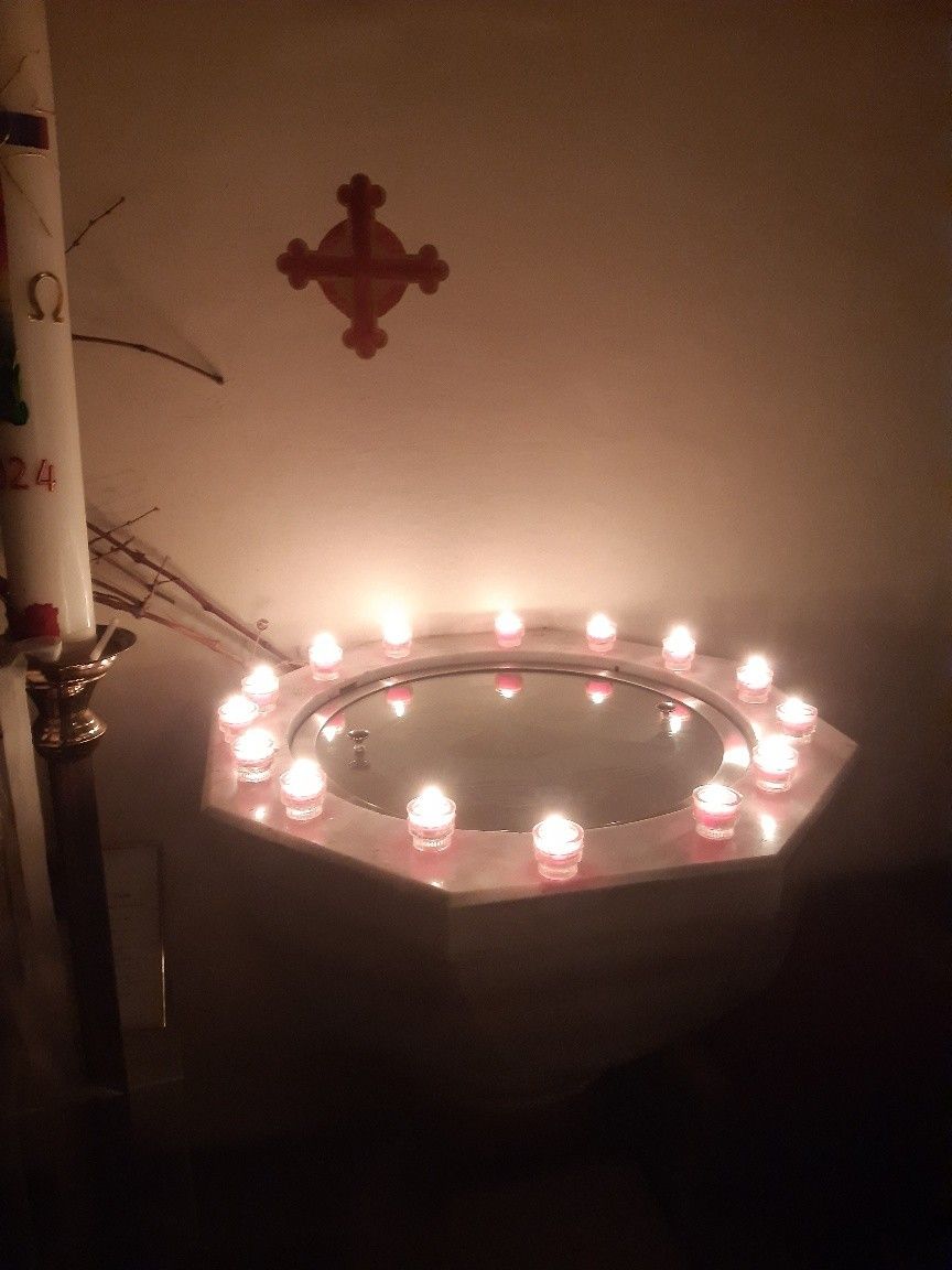 Ein schwach beleuchteter Raum zeigt eine kreisförmige Anordnung von Kerzen um einen reflektierenden Pool, mit einem Kreuz darüber und einem Kerzenständer in der Nähe.