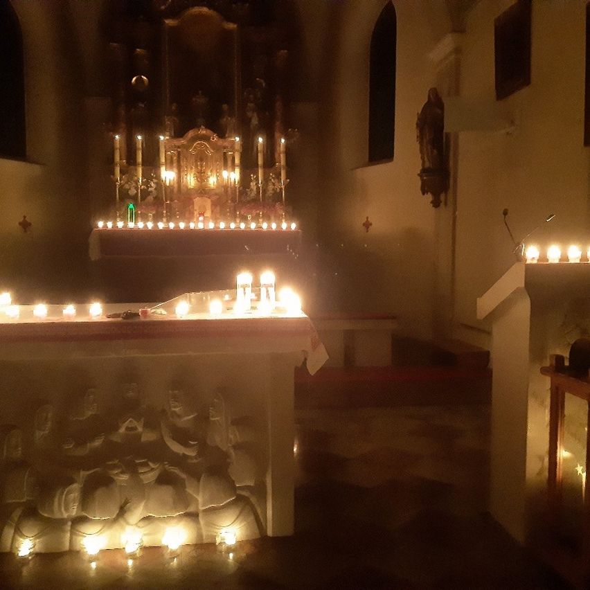 Ein schwach beleuchteter Raum mit Kerzen und Statuen, einer geschnitzten Szene auf dem Altar und einer an der Wand montierten Statue.