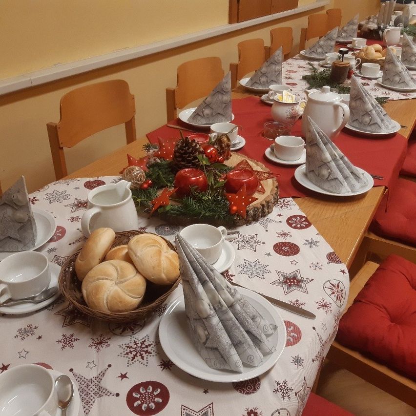 Ein langer Esstisch für das Frühstück mit Brot, Kaffeetassen und einem Weihnachts-Mittelstück. Der Tisch ist mit einem roten Tuch und einer gemusterten Tischdecke bedeckt.