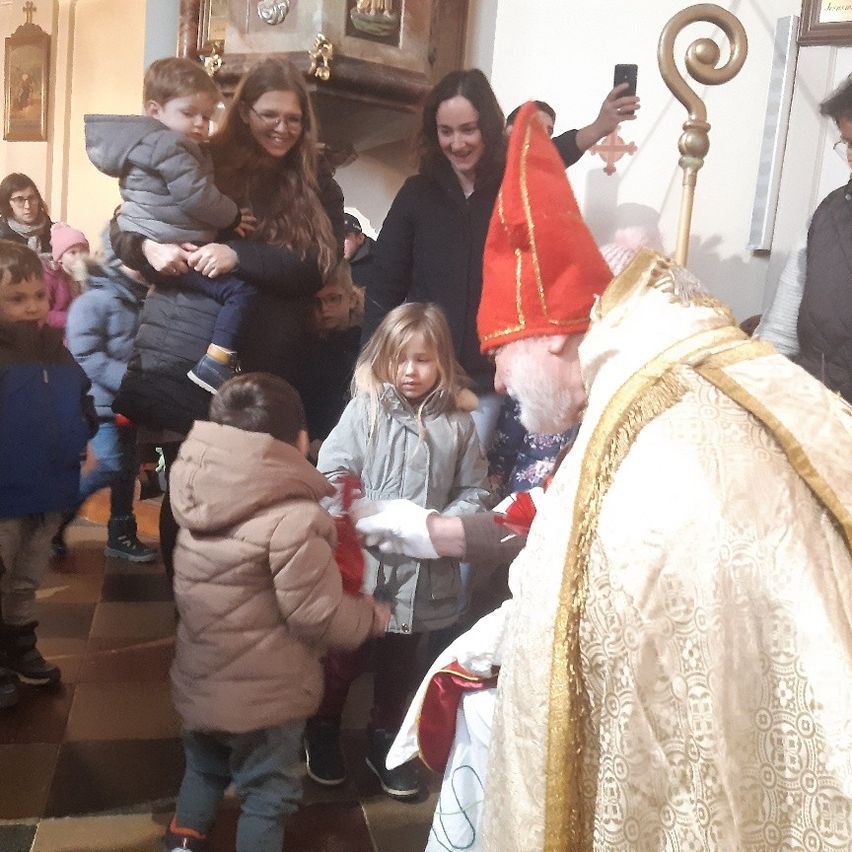 Ein Priester in roten Gewändern segnet Kinder in einer Kirche. Erwachsene und Kinder sind versammelt, einige machen Fotos. Der Priester hält ein rotes Objekt, möglicherweise ein Buch.