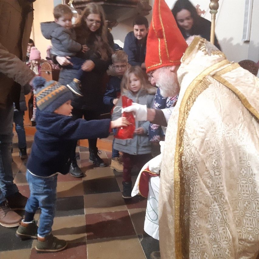 Ein Priester gibt einem kleinen Kind während einer Kirchenzeremonie ein Geschenk. Andere Kinder und Erwachsene beobachten.