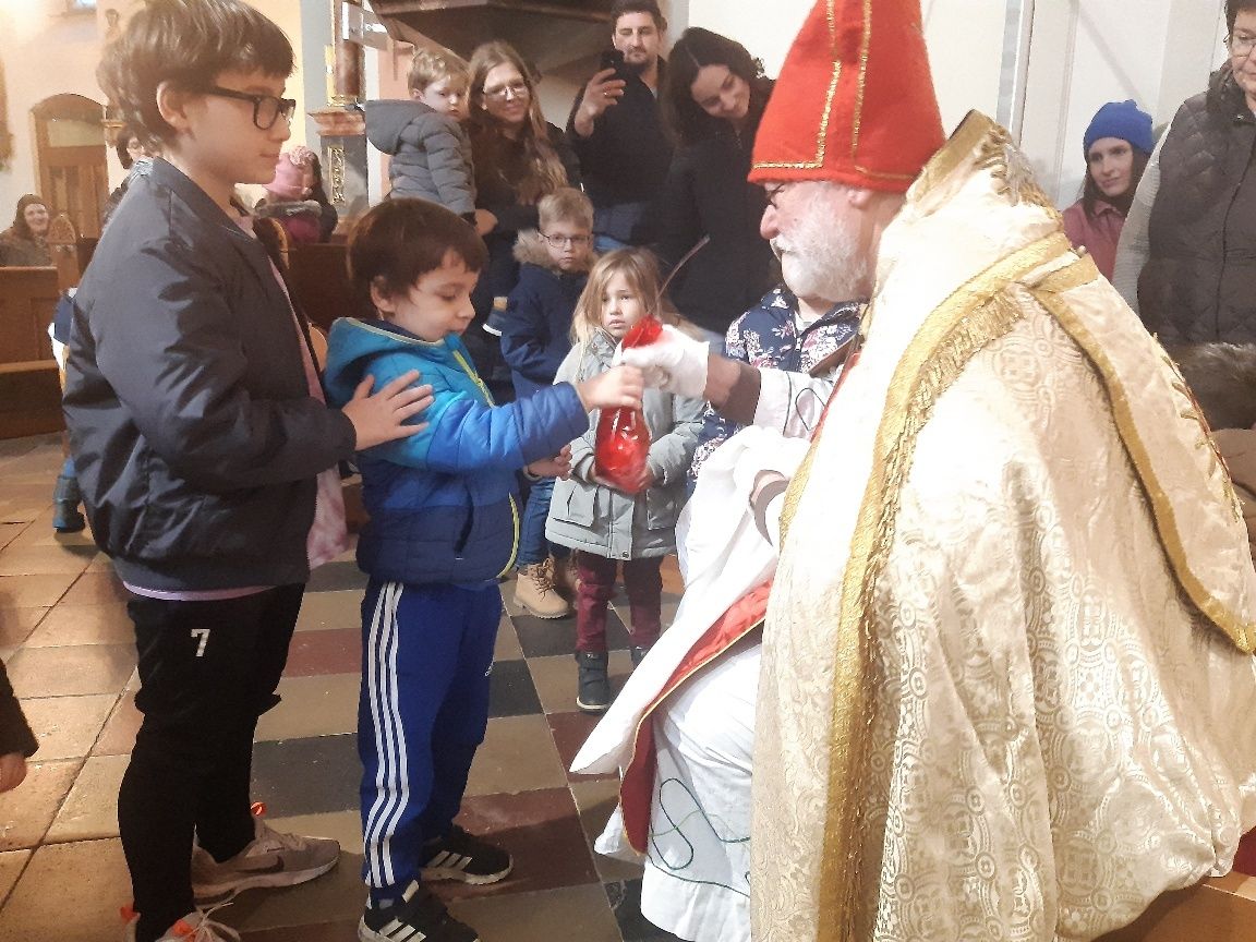 Ein Priester in traditioneller Kleidung reicht einem Kind in einer blauen Jacke einen roten Gegenstand, während andere Kinder und Erwachsene zusehen.
