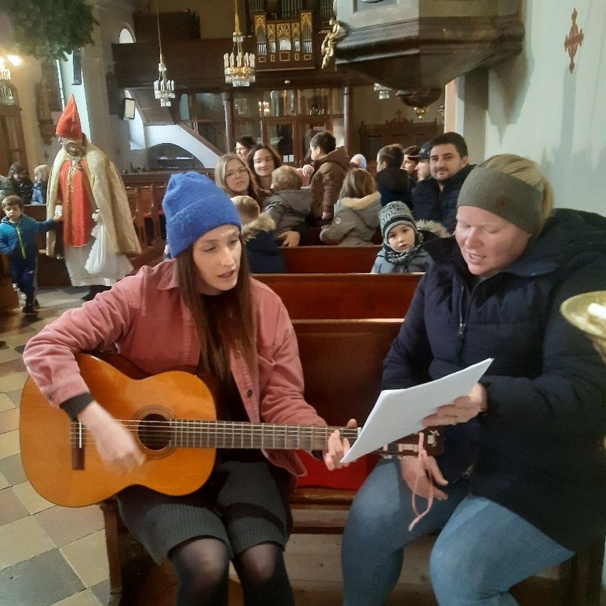Eine Frau spielt Gitarre in einer Kirche, während eine andere von einem Stück Papier liest; Menschen sitzen hinter ihnen auf den Kirchenbänken.