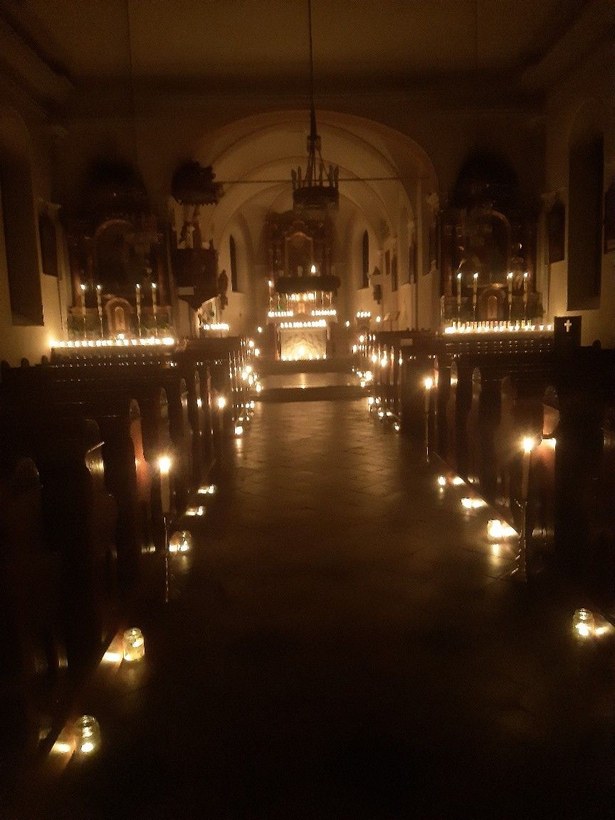Eine schwach beleuchtete Kirche mit Bankreihen und Kerzen, die den Gang säumen, zum großen Altar mit vielen brennenden Kerzen.