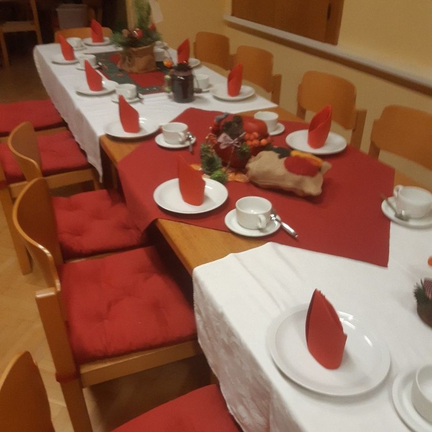 Ein Esstisch ist mit roten Tischdecken und weißen Tellern, Tassen und Servietten gedeckt. Dekorative Gegenstände wie eine Kürbis und ein Korb stehen auf dem Tisch. Holzstühle mit roten Kissen umgeben den Tisch.