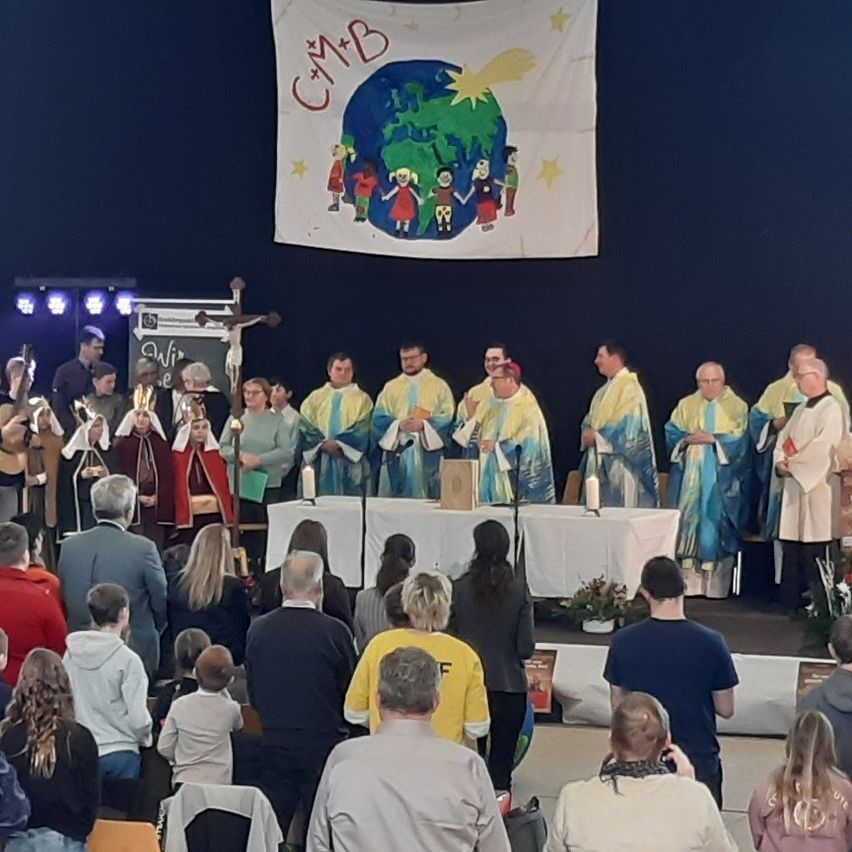 Eine religiöse Zeremonie mit einem Banner, einem Kreuz und einer Gruppe von Menschen in Gewändern, die auf einer Bühne mit Tischen und Kerzen stehen.